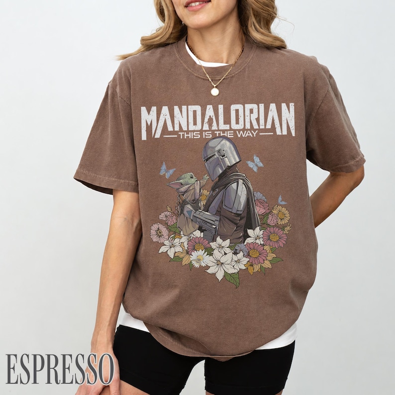 May include: Espresso-coloured t-shirt with the text "MANDALORIAN - THIS IS THE WAY" above an illustration of the Mandalorian and Grogu, surrounded by flowers and butterflies. The shirt has a relaxed fit and a white undershirt.