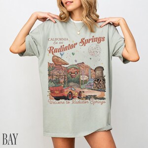 May include: A sage green short-sleeve t-shirt featuring a graphic print of Radiator Springs with a red race car, desert rock formations, and a water tower. The text reads California Est. 2012, Radiator Springs, and Welcome to Radiator Springs.