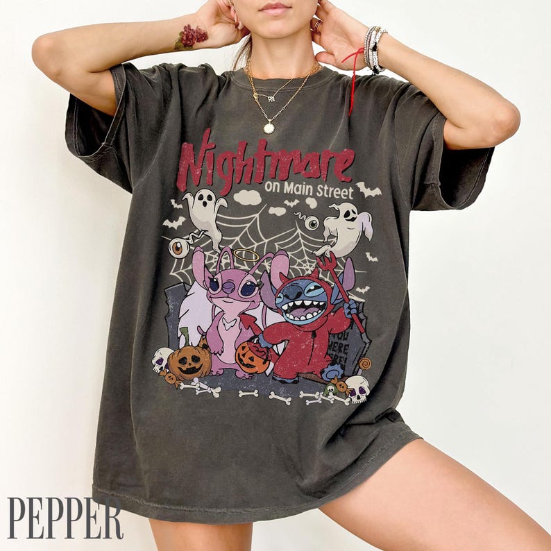 May include: A charcoal gray short-sleeve t-shirt featuring a graphic print with the text Nightmare on Main Street. The design includes two blue and pink alien characters, ghosts, spiderwebs, pumpkins, and skulls in a Halloween theme.