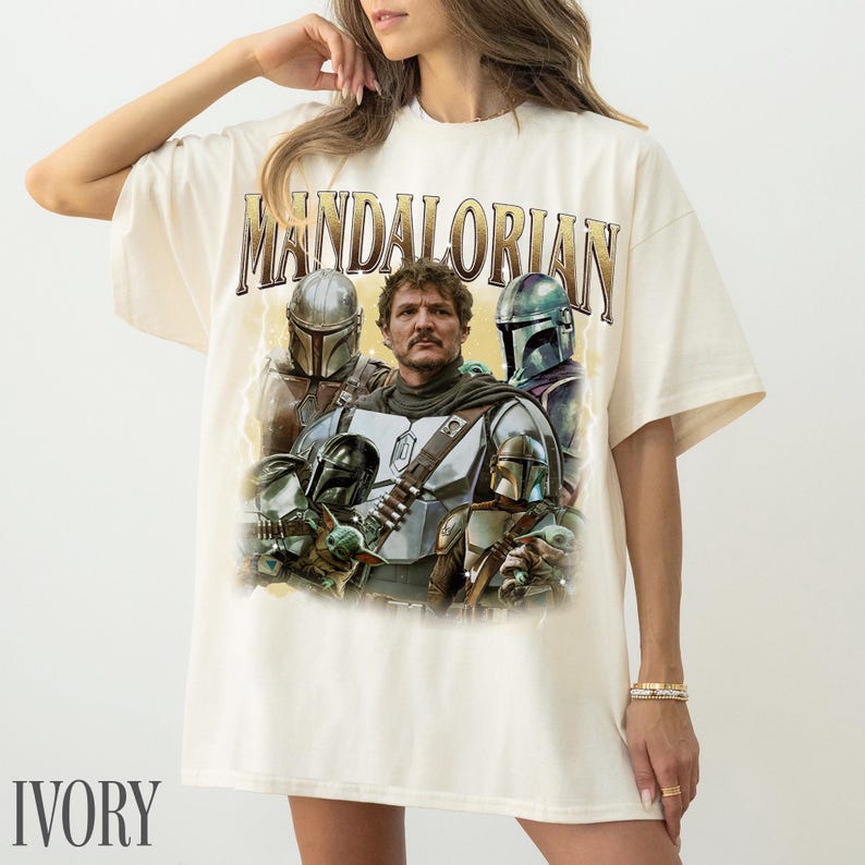 May include: Ivory t-shirt with the word "MANDALORIAN" in gold lettering. The graphic features characters from the series. The shirt has a loose fit.