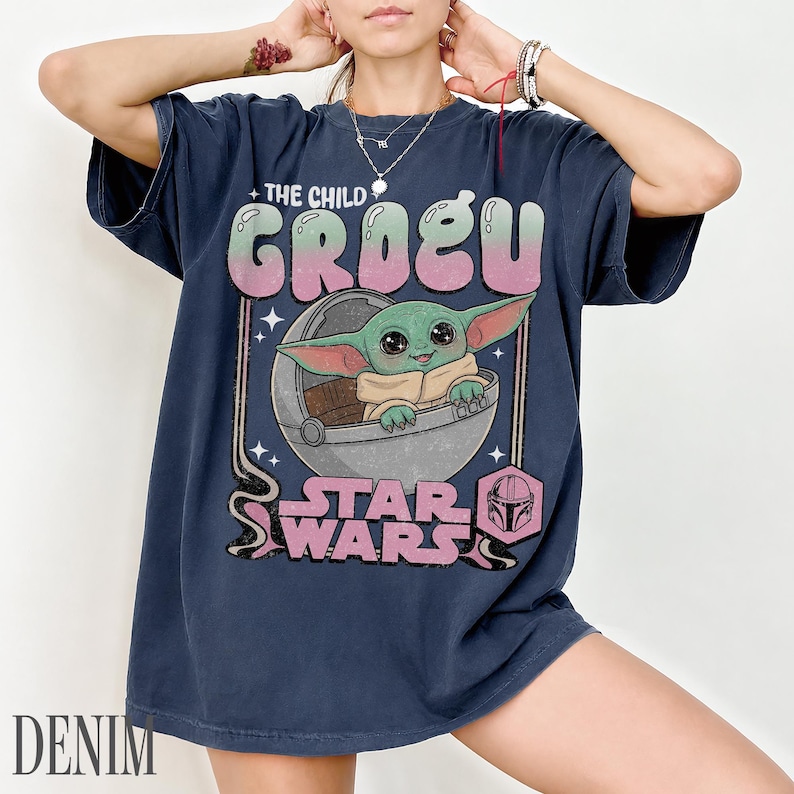 May include: A dark blue short-sleeve t-shirt featuring a graphic of Grogu from Star Wars sitting in a floating pod. The text reads THE CHILD GROGU and STAR WARS in a retro pink and green font. The design includes small star accents and a helmet icon.
