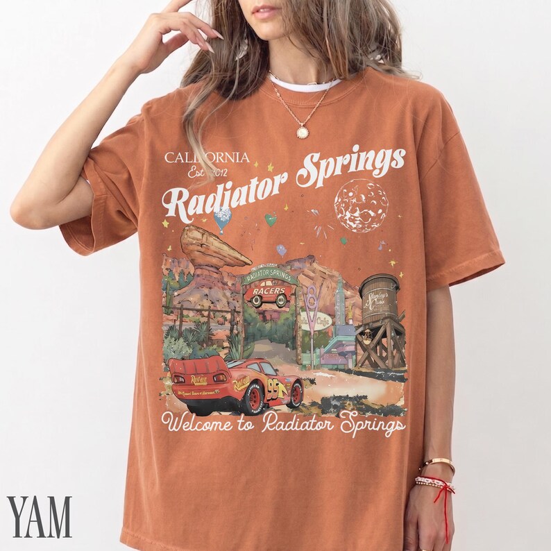 May include: A rust-coloured short-sleeve t-shirt featuring a graphic print of Radiator Springs with a red race car, desert rock formations, a water tower, and the text California Est 2012 Radiator Springs Welcome to Radiator Springs.