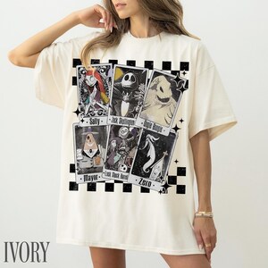 May include: Ivory-colored t-shirt featuring a graphic of tarot-style cards with characters from The Nightmare Before Christmas. The cards include Sally, Jack Skellington, Oogie Boogie, Mayor, Lock Shock Barrel, and Zero. The shirt has a black and white checkered pattern.