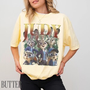May include: Yellow t-shirt featuring a graphic of Judy Hopps from Zootopia. The design includes the word "JUDY" in gold lettering and images of Judy Hopps in various poses. The word "BUTTER" is printed in the bottom left corner.