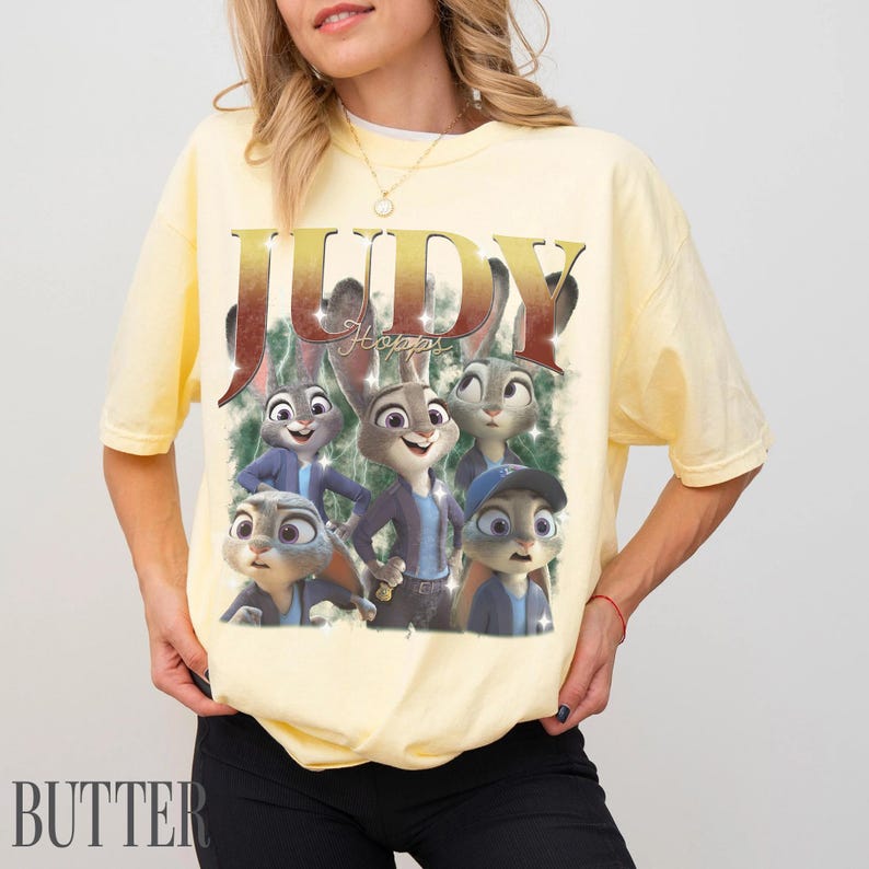 May include: Yellow t-shirt featuring a graphic of Judy Hopps from Zootopia. The design includes the word "JUDY" in gold lettering and images of Judy Hopps in various poses. The word "BUTTER" is printed in the bottom left corner.