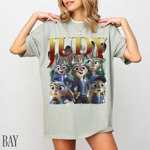 May include: A light green t-shirt featuring a graphic of Judy Hopps from Zootopia. The design includes the name "JUDY" in gold letters and multiple images of the character. The shirt has short sleeves and a relaxed fit.