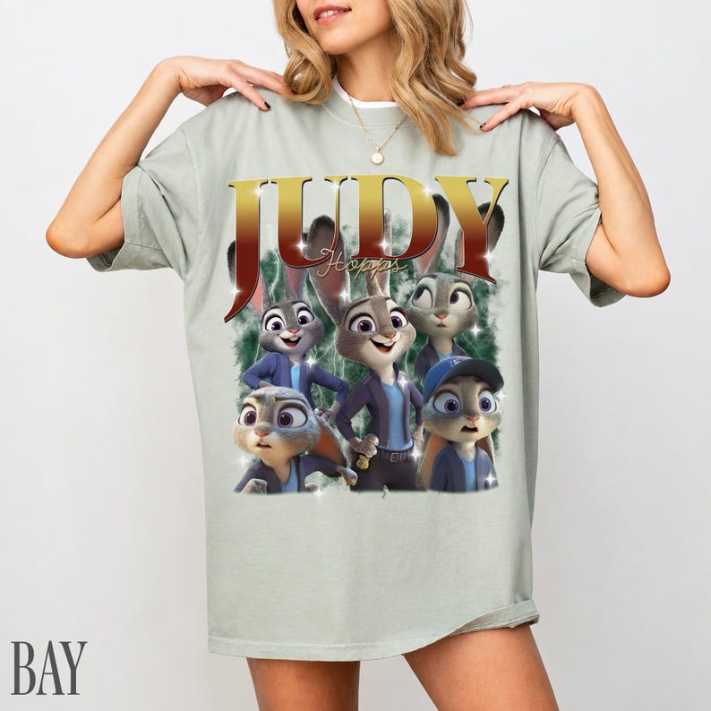 May include: A light green t-shirt featuring a graphic of Judy Hopps from Zootopia. The design includes the name "JUDY" in gold letters and multiple images of the character. The shirt has short sleeves and a relaxed fit.