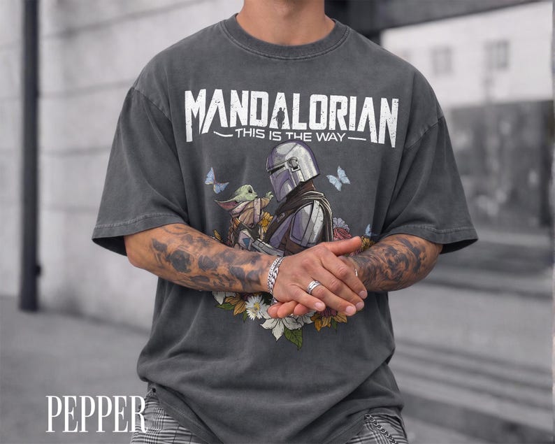May include: Charcoal grey t-shirt with the white text "MANDALORIAN THIS IS THE WAY". The design shows the Mandalorian and Grogu with flowers and butterflies. The word "PEPPER" is printed at the bottom of the shirt.