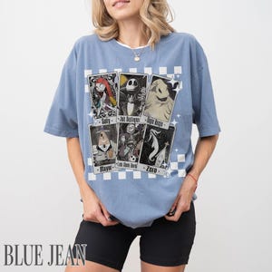 May include: A light blue t-shirt featuring a graphic of playing cards with characters from The Nightmare Before Christmas. The cards include Sally, Jack Skellington, Oogie Boogie, Mayor, Lock, Shock, Barrel, and Zero. The words "BLUE JEAN" are printed at the bottom.