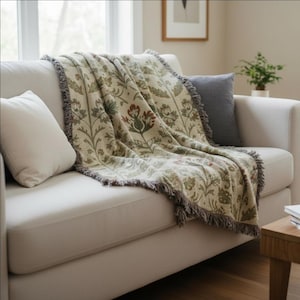 Puede incluir: Una manta beige con estampado floral y detalles en verde y rojo, colocada sobre un sofá blanco. La manta tiene un borde con flecos y se combina con un cojín gris y un cojín blanco. Una pequeña planta es visible en el fondo.