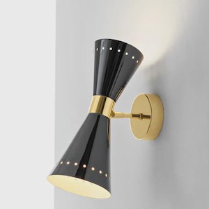 Peut inclure: Applique murale noire et dorée de style moderne du milieu du siècle. L'applique présente deux abat-jours coniques avec de petits trous espacés, une bande dorée et une plaque de montage dorée. Le luminaire est fixé sur un mur gris clair.