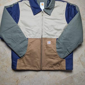 May include: A colour-blocked zip-up jacket featuring a beige upper chest, brown lower front panels, and olive green sleeves with navy blue side accents. The jacket has a standard collar, a central metal zip, and a Carhartt brand label on the chest.