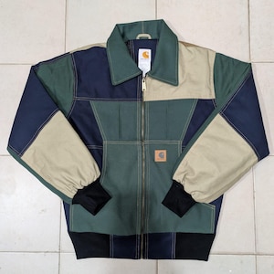 Peut inclure: Une veste Carhartt à blocs de couleurs composée de panneaux vert foncé, bleu marine et beige. La veste présente une fermeture éclair intégrale, un col pointu, des manches longues avec des poignets côtelés noirs et un ourlet côtelé noir.