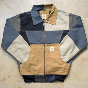 May include: A colour-blocked zip-up jacket featuring a patchwork design in shades of tan, charcoal grey, and black. The jacket has a classic collar, a full-length metal zip, and a Carhartt brand label on the chest pocket and inner neck.