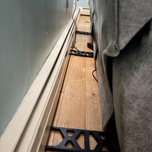 May include: Black metal furniture supports are placed on a hardwood floor between a wall and a gray upholstered sofa. Electrical cords run along the wall and floor. The wall is painted a light blue-green color, and the baseboard is off-white.
