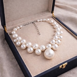 May include: A pearl necklace with a large central pearl, displayed in a dark blue jewelry box with a beige interior. The necklace features multiple sizes of round, white pearls, creating a clustered design.