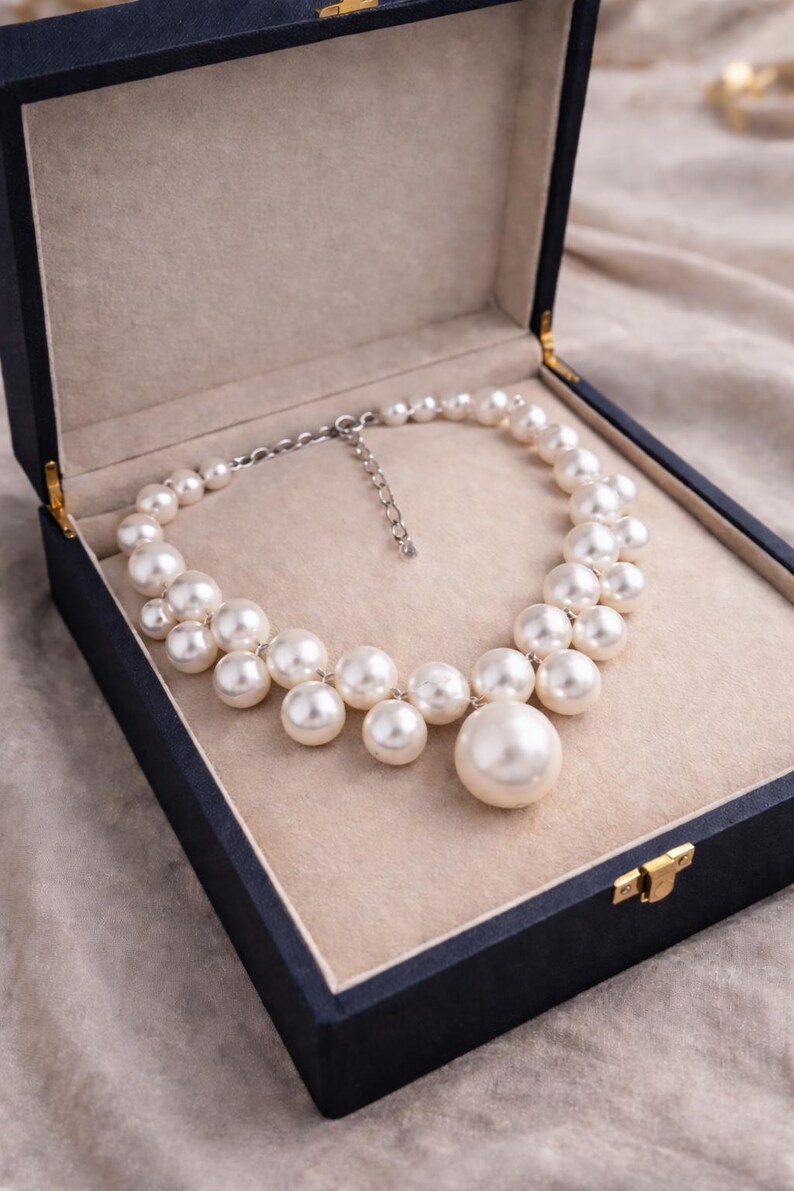 May include: A pearl necklace with a large central pearl, displayed in a dark blue jewelry box with a beige interior. The necklace features multiple sizes of round, white pearls, creating a clustered design.