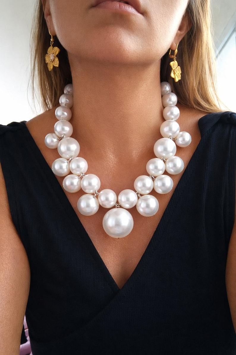 May include: A statement pearl necklace with large, round, white pearls of varying sizes. The necklace is paired with gold-colored flower earrings. The model is wearing a black top.