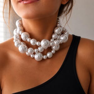 Peut inclure: Un collier imposant à plusieurs rangs composé de perles artificielles blanches brillantes de tailles variées. Les perles sont disposées en un motif superposé et groupé qui repose près du cou, créant un aspect volumineux et texturé.