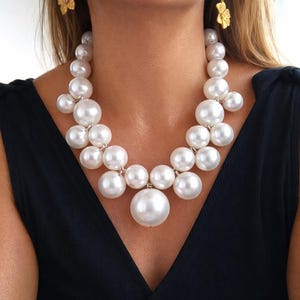 May include: A striking pearl necklace featuring large, round, white pearls of different sizes. The necklace is complemented by gold-coloured flower earrings. The model is wearing a black top.