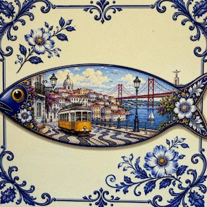 Könnte beinhalten: Eine dekorative Keramikfliese in Fischform mit einer detaillierten Darstellung einer gelben Straßenbahn auf einer gepflasterten Straße in Lissabon, Portugal. Die Szene zeigt die Brücke des 25. April, ein Stadtbild und blaue Blumenmuster.