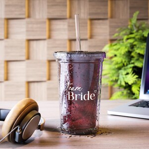 May include: A clear plastic tumbler filled with a dark red liquid, featuring the text "Team Bride" in white script. The tumbler has a black lid and a clear straw. The image is set on a wooden surface.