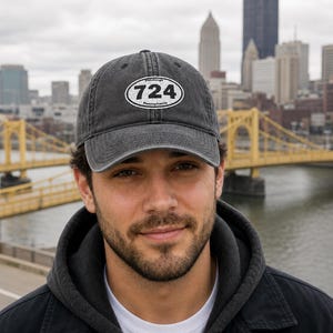 May include: A gray baseball cap with the text "Pittsburgh 724 Pennsylvania" in a white oval. The cap is worn by a person in front of a city skyline with a yellow bridge.