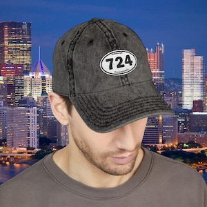 May include: A charcoal grey baseball cap with a distressed finish. The cap has a white oval patch with the number "724" and the text "Pittsburgh Pennsylvania". The background shows a cityscape at dusk.