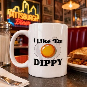 May include: White ceramic mug with the text "I Like 'Em Dippy" and a graphic of a fried egg. The mug is on a table with a diner setting in the background.