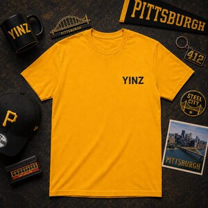 May include: A gold short-sleeved t-shirt with the word YINZ printed in black on the chest. The shirt is surrounded by Pittsburgh-themed items including a black mug, a baseball cap with a P, a bridge model, a pennant, a keychain, and a photograph.