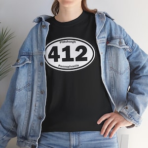 May include: Black t-shirt featuring a white oval design with the text "Pittsburgh" above the large numbers "412" and "Pennsylvania" below. The shirt is worn with a denim jacket.