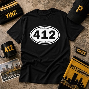 May include: A black t-shirt featuring a white oval graphic with the text 412, Pittsburgh, and Pennsylvania. Surrounding items include a black and yellow beanie with YINZ, a black baseball cap with a P, a black mug with 412, a keychain with YINZER, a photo of the Pittsburgh skyline, and a yellow and black towel with the word Pittsburgh.