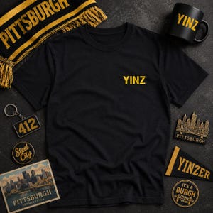 May include: A black short-sleeved t-shirt featuring the yellow text YINZ printed on the chest. The shirt is surrounded by Pittsburgh-themed items including a black and yellow scarf, a black mug with YINZ text, a 412 keychain, and city-themed decals.