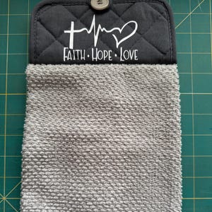 May include: Gray kitchen towel with a black quilted top featuring the words "Faith Hope Love" in white, along with a cross, heartbeat, and heart symbol. The towel has a loop and button closure.