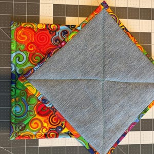 May include: A square, quilted pot holder with a denim gray interior and a colorful, swirling pattern on the exterior. The edges are trimmed with a patterned fabric in shades of red, orange, yellow, green, blue, and purple.
