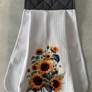 May include: White kitchen towel with a sunflower design. The towel has a gray quilted top with a button closure. The sunflower design features yellow and orange blooms with green leaves.