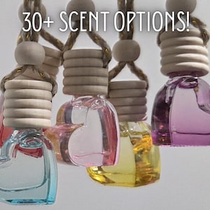 Puede incluir: Varios ambientadores de vidrio en forma de corazón en varios colores, incluyendo azul, rosa y amarillo, están suspendidos de una cuerda. El texto "30+ SCENT OPTIONS!" está visible. Cada botella tiene una tapa de madera.
