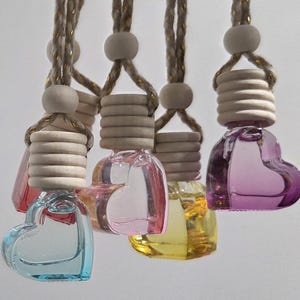 Puede incluir: Ambientadores para coche con forma de corazón de cristal en varios colores, como azul, rosa, amarillo y morado. Cada botella tiene una tapa de madera y un cordón trenzado para colgar. Las botellas están llenas de líquido coloreado.