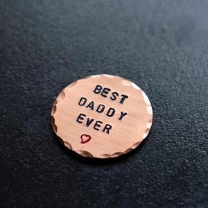 Op de afbeelding: Een ronde koperen penning met een gehamerde rand, voorzien van de gestempelde zwarte tekst BEST DADDY EVER en een klein rood hartsymbool onder de tekst. Het metalen oppervlak heeft een geborstelde afwerking en een duidelijke koperkleur.