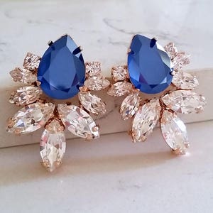 May include: Rose gold earrings with a large blue teardrop-shaped crystal surrounded by smaller clear crystals.