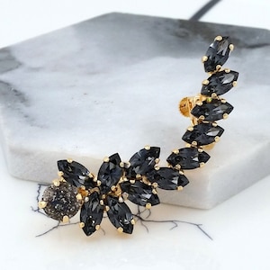 May include: A gold ear cuff with a cluster of black marquise-shaped crystals. The ear cuff is designed to sit on the upper part of the ear.