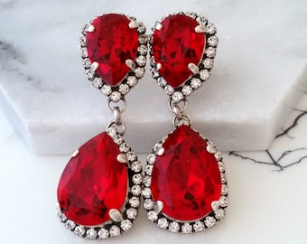 May include: A pair of dangle earrings with two teardrop-shaped red crystals. Each crystal is surrounded by a halo of smaller clear crystals.