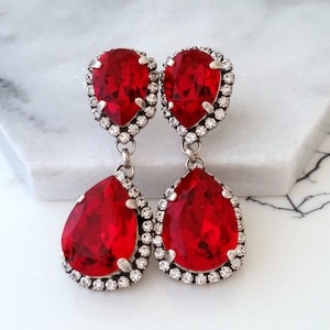 以下が含まれることがあります： 2 つの涙型の赤いクリスタルが付いた、ぶら下がっているイヤリングのペア。各クリスタルは、より小さな透明なクリスタルのハローで囲まれています。