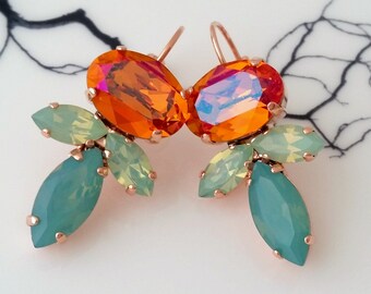 May include: A pair of rose gold earrings featuring large, oval orange crystals with iridescent highlights. The earrings also have smaller, leaf-shaped crystals in shades of green and teal. The design is set against a white background.