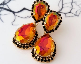 May include: A pair of gold tone dangle earrings with two teardrop shaped iridescent orange crystals. The crystals are surrounded by a black rhinestone border.