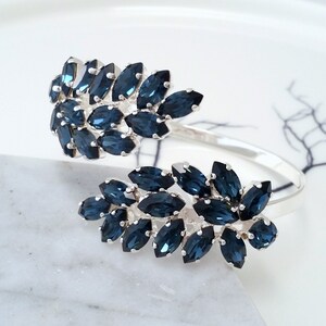 May include: A silver bangle bracelet with two clusters of dark blue marquise-shaped rhinestones. The rhinestones are set in a leaf-like pattern.