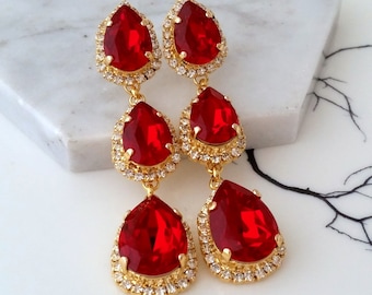 May include: A pair of long, dangling earrings with three teardrop-shaped red crystals. Each crystal is surrounded by a gold-toned setting with smaller clear crystals.