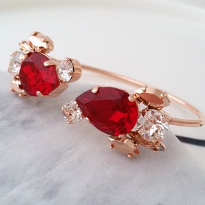 May include: A gold bangle bracelet featuring red and clear crystal embellishments. The bracelet is open and has a delicate design.