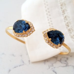 May include: A gold bangle bracelet with two teardrop-shaped blue gemstones surrounded by smaller clear rhinestones.