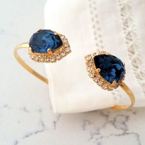 May include: Gold bangle bracelet with two teardrop-shaped blue gemstones surrounded by clear rhinestones.